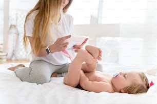 Cheerful young woman is changing diaper of her little daughter. She is sitting on bed and smiling