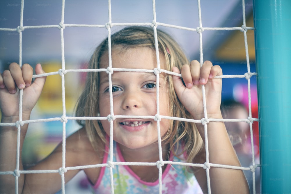 Menina sorridente pequena e bonito sentada na cerca protetora. Menina brincando no playground.  Olhando para a câmera.