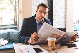 Serious stubbled businessman looking at newspaper while locating at desk in room