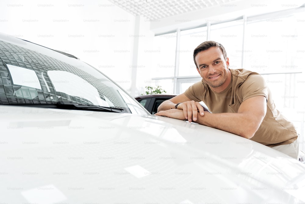 Retrato de hombre demostrando felicidad mientras se apoya en el capó blanco en el concesionario de automóviles