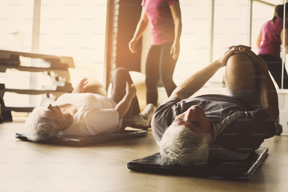 Treino de casal sénior em centro de reabilitação. Personal trainer ajuda casal de idosos a fazer alongamento no chão." n