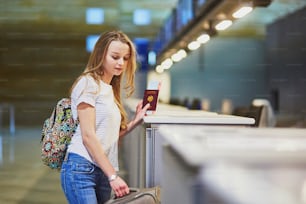 Beautiful young tourist girl with backpack and carry on luggage in international airport at check-in counter, giving her passport to an officer and waiting for her boarding pass