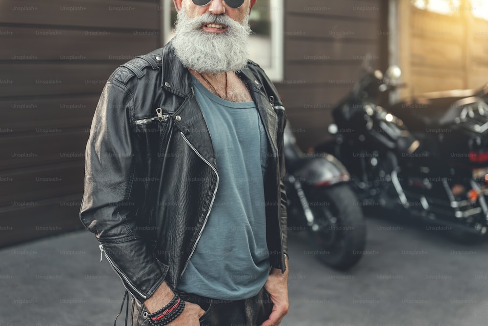 Esilarante vecchio motociclista barbuto con gli occhiali indossa la pelle. Lui in piedi vicino al garage con il veicolo. Ritratto