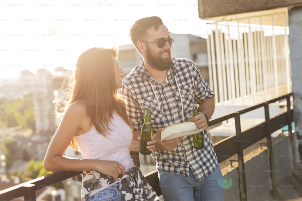Young couple having fun at a rooftop party, drinking beer, eating barbecue and chatting
