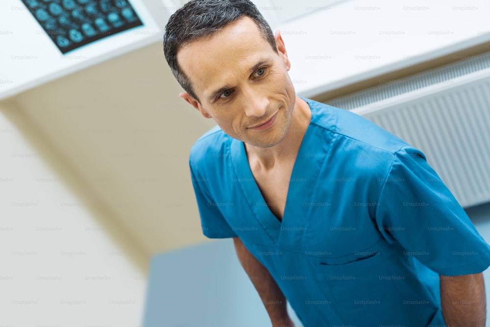 Trabajador agradable. Hombre guapo de mediana edad con un uniforme azul de enfermero masculino de pie en el laboratorio de tomografía computarizada, mirando a lo lejos y sonriendo