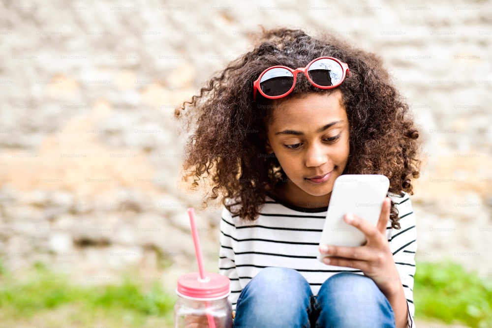 Belle fille afro-américaine aux cheveux bouclés à l’extérieur portant un t-shirt rayé tenant un téléphone intelligent, lisant quelque chose ou envoyant des SMS, buvant de l’eau dans un verre avec de la paille rose.