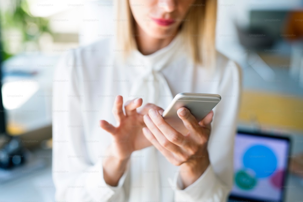 Femme d’affaires méconnaissable dans son bureau tenant un téléphone intelligent, envoyant des SMS.