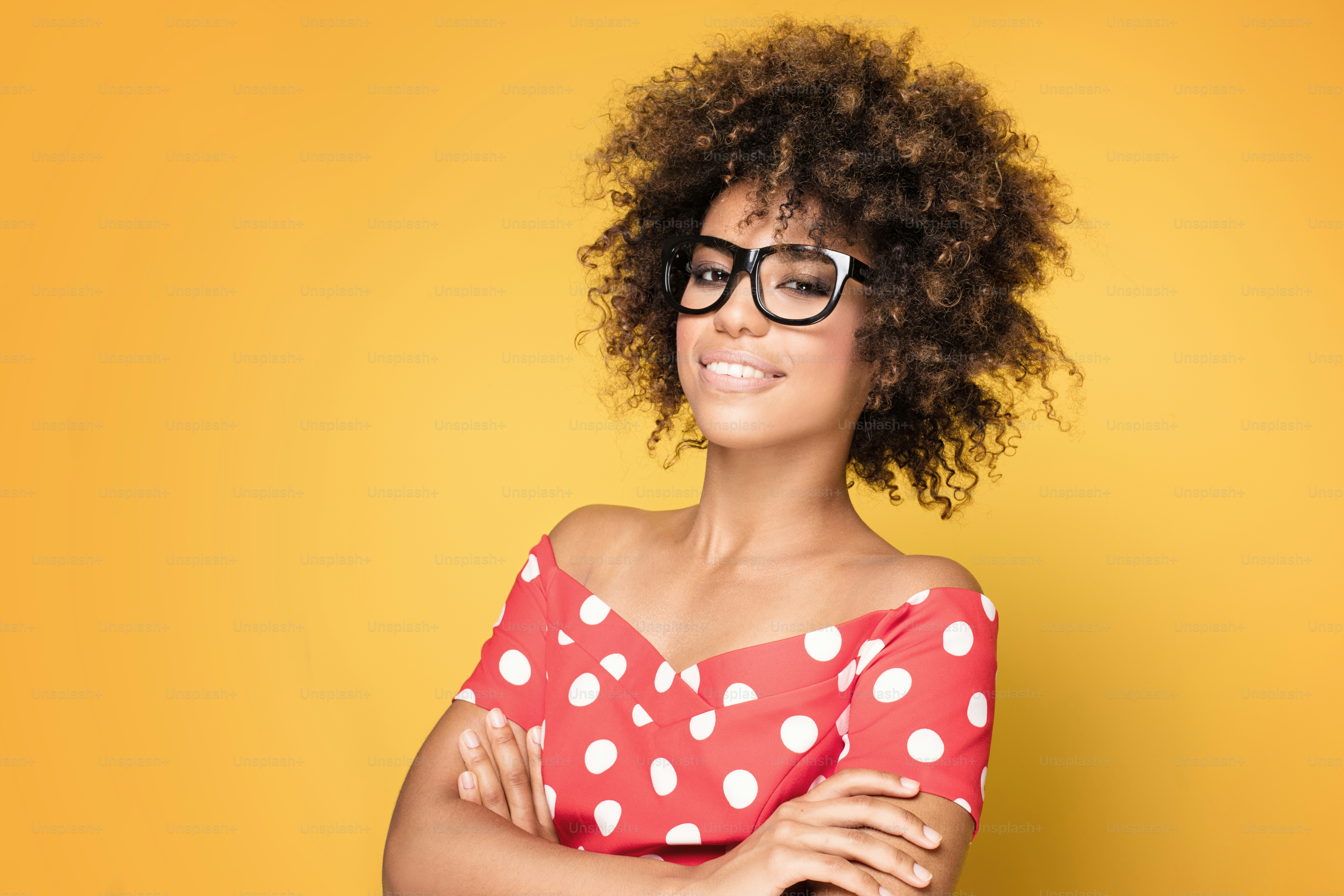 Retrato de una hermosa mujer joven afroamericana sonriente. Chica con afro con gafas. Fondo amarillo. Toma de estudio.
