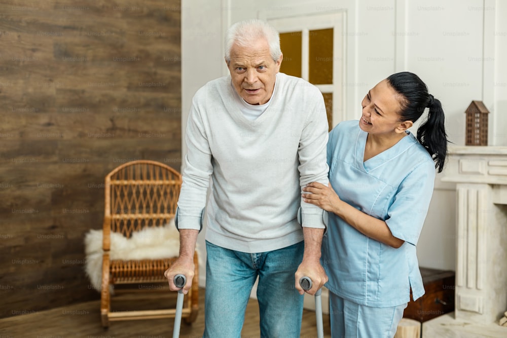 Asistencia médica. Buen hombre de edad avanzada positivo que usa bastón y camina en la habitación mientras recibe el apoyo de una cuidadora profesional