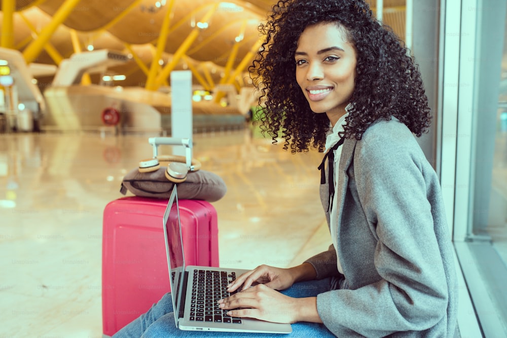 Mujer negra que trabaja con una computadora portátil en el aeropuerto esperando en la ventana