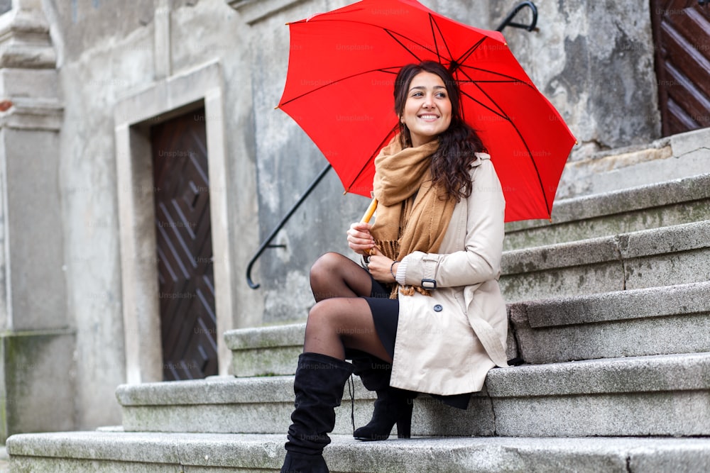 Joven sonriente con paraguas rojo sentada en las escaleras del casco antiguo.