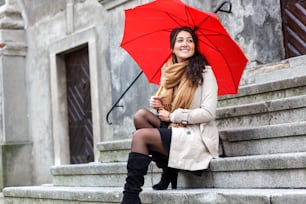 Smiling young woman with red umbrella sitting on the stairs in the old town.