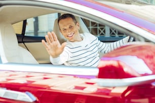 Professional driver. Cheerful nice positive man sitting behind the wheel and smiling while greeting his friend