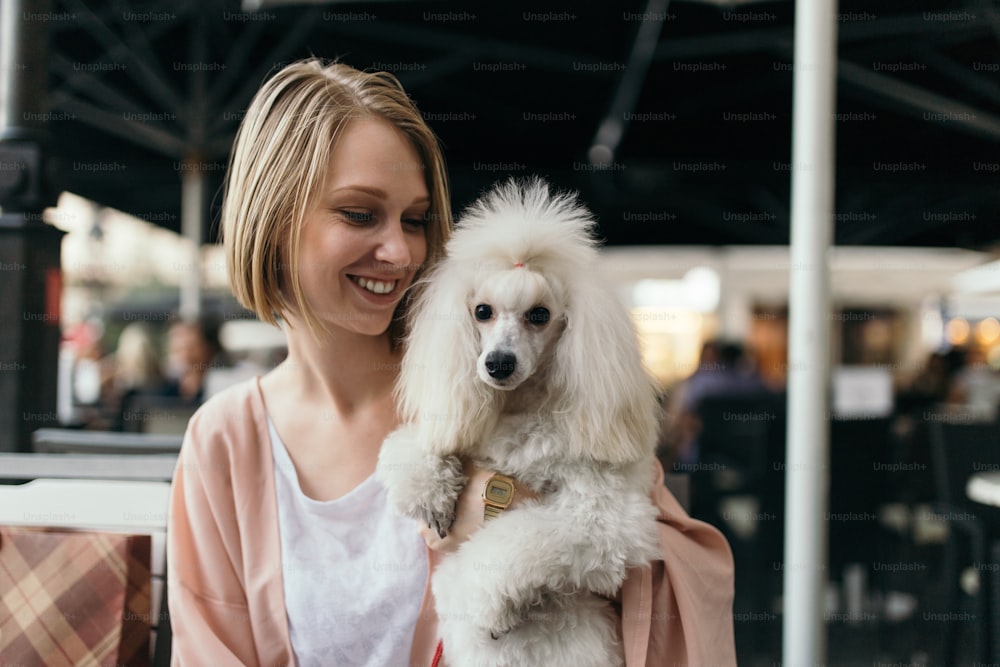 Beautiful and fashionable young woman sitting in cafe bar with her gorgeous white dwarf poodle and looking at camera