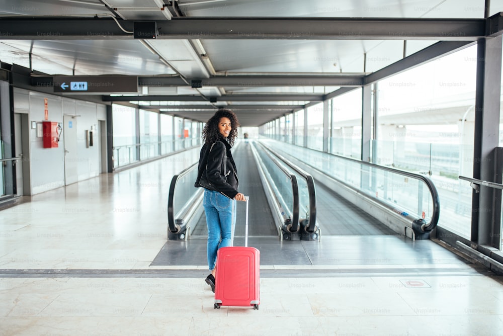 Mujer negra en la pasarela móvil del aeropuerto con una maleta rosa.