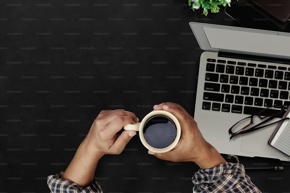Mani dell'uomo che tengono la pausa caffè mentre lavorano sulla tastiera del computer portatile con monitor a schermo vuoto sul tavolo in pelle con superficie scura.