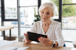 Dispositivo moderno. Empresária profissional encantada e inteligente sentada à mesa e usando um tablet enquanto trabalha no escritório