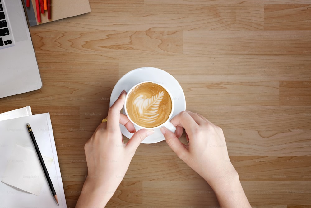 モダンな木製のオフィスデスクと、カフェラテアートコーヒーのカップを持つ女性。