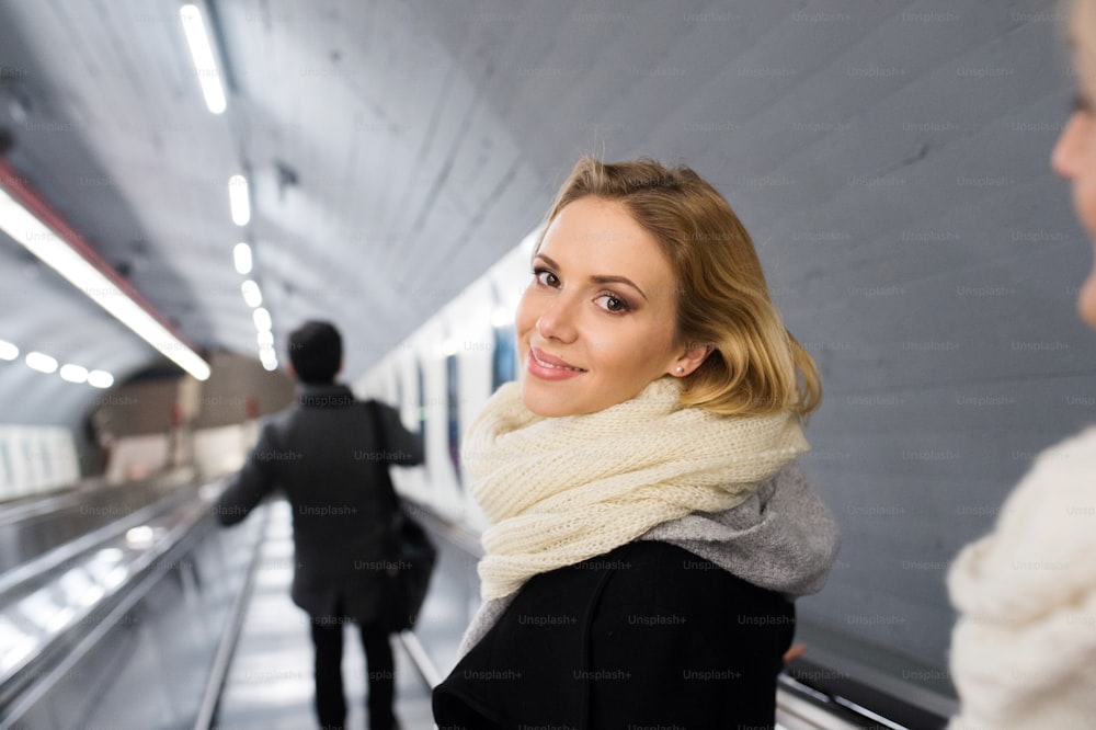 검은 코트와 큰 모직 스카프를 입은 아름다운 젊은 금발 여자가 비엔나 지하철의 에스컬레이터에 서 있다