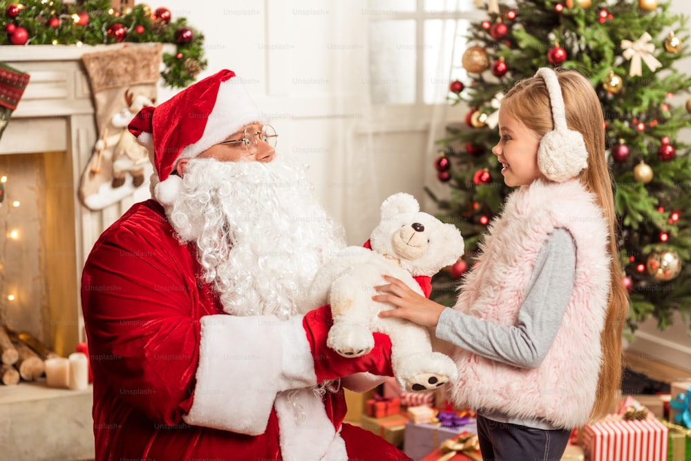 This present is for you. Kind senior bearded man in red and white costume is giving teddy bear to child. Girl is standing near Christmas tree and laughing