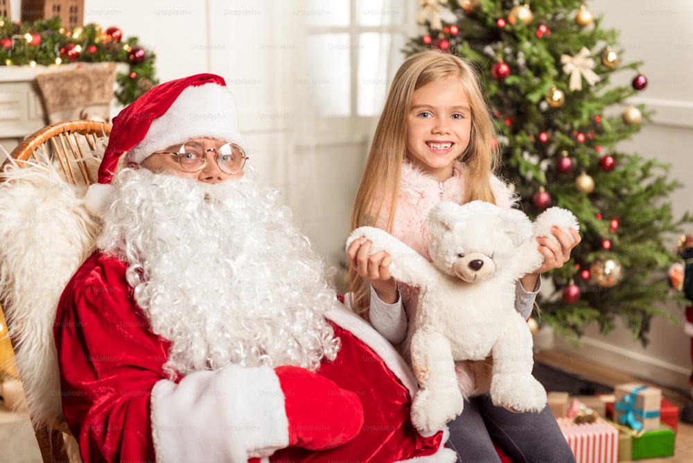 Christmas is my favorite holiday. Happy little girl is sitting on knees of Santa Claus and holding teddy bear. She is looking at camera and smiling