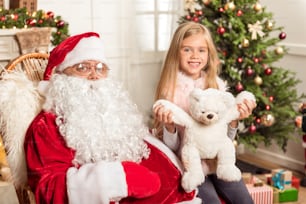 Christmas is my favorite holiday. Happy little girl is sitting on knees of Santa Claus and holding teddy bear. She is looking at camera and smiling