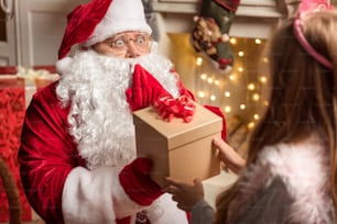 Ti meriti questo regalo. Ritratto di Babbo Natale eccitato che dà scatola alla bambina. È seduto su una comoda poltrona in costume rosso