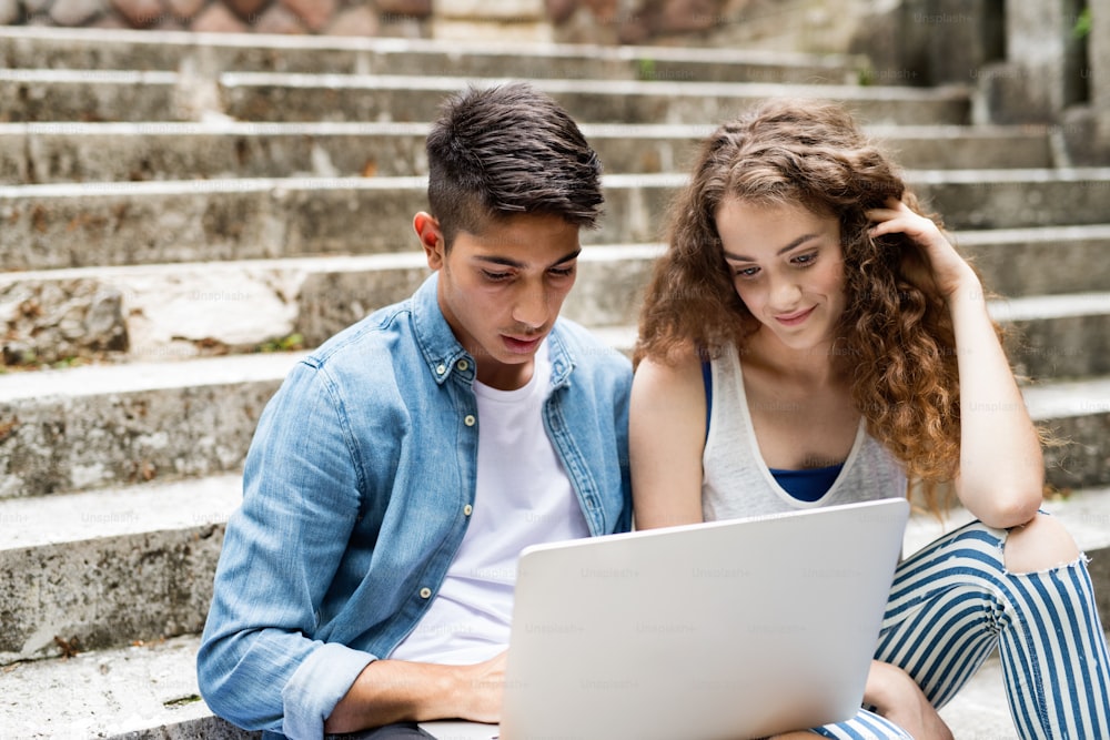 Attractive teenage student couple sitting on stone steps in front of university with laptop, reading or watching something.
