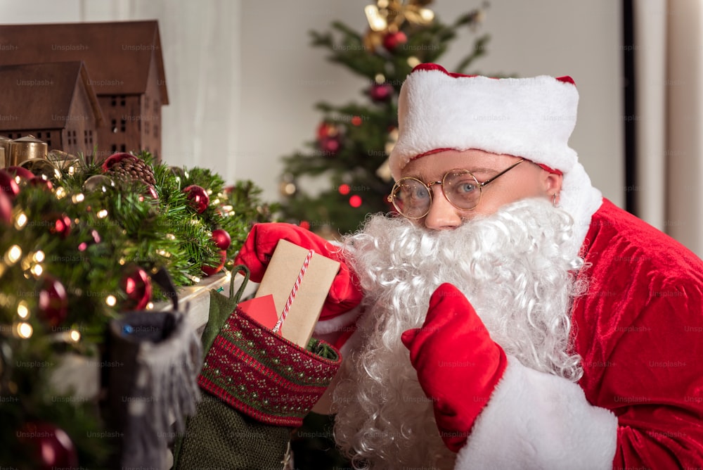 Porträt des mysteriösen Weihnachtsmannes, der heimlich gestikuliert, während er eine Geschenkbox in einer großen dekorativen Socke versteckt. Er schaut mit Freude in die Kamera