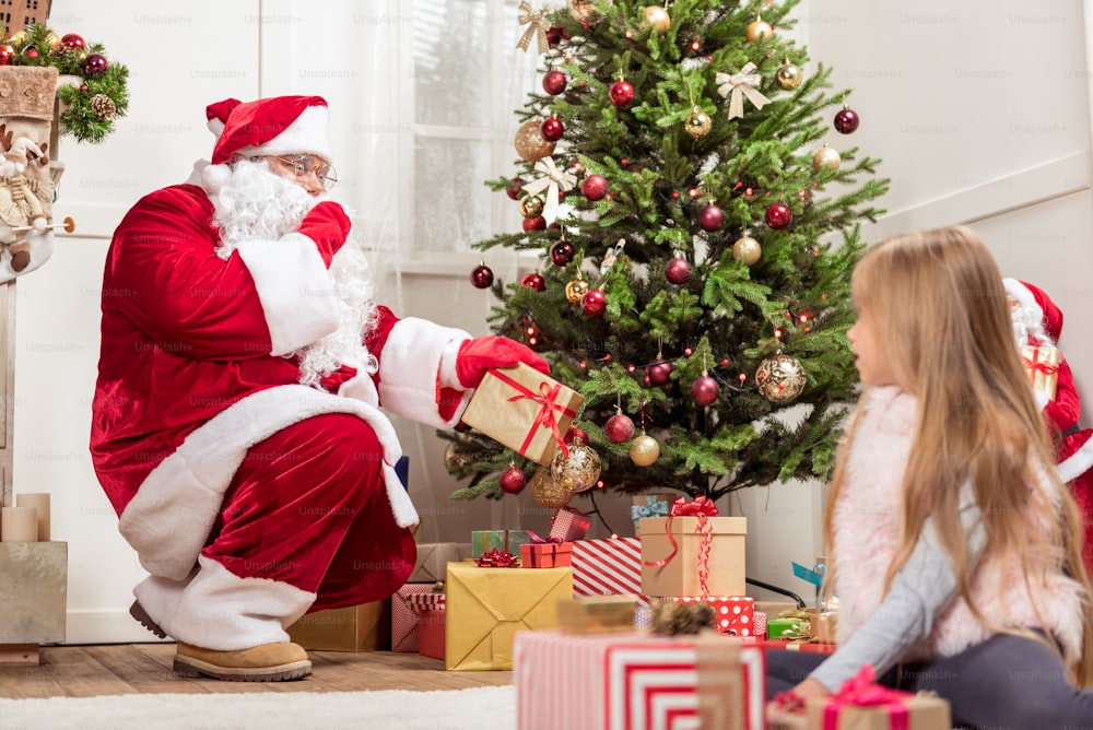 Hush-hush. Mysterious Santa Claus is laying present boxes under Christmas tree. He is gesturing secretly to curious girl, who is sitting on floor