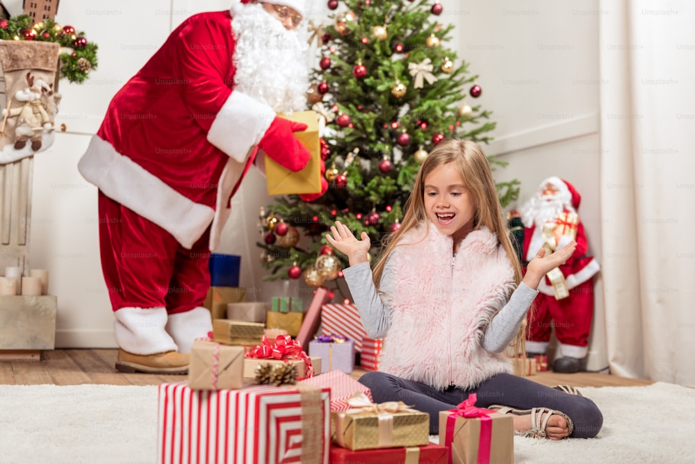 Portrait of joyful girl looking at many present boxes with excitement. She is sitting on floor and laughing. Santa Claus is putting presents under the Christmas tree on background