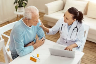 Dont worry. Cheerful friendly positive doctor looking at her senior patient and comforting him while working on the laptop