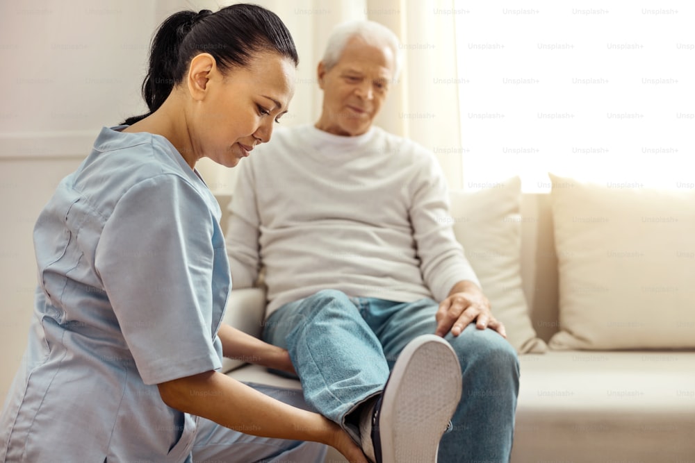 Rehabilitation therapy. Delighted pleasant professional nurse smiling and looking at her aged patients leg while holding it