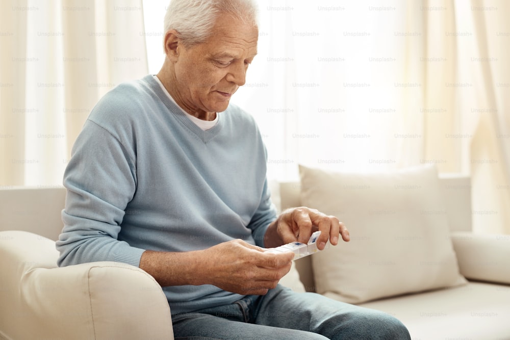 Prescrição médica. Alegre homem idoso agradável segurando um organizador de pílulas e tomando seu remédio enquanto está sentado no sofá