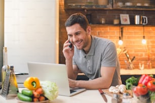 Young man vegan using laptop and talking phone while preparing dinner