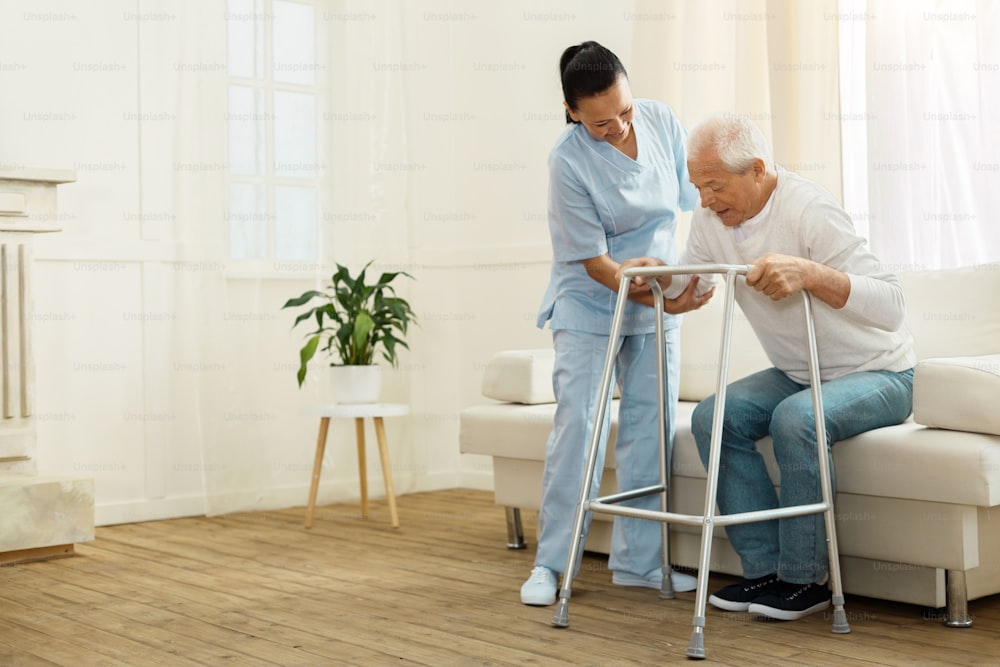 Do not hurry. Delighted positive cheerful caregiver smiling and helping her patient to stand up while being near him