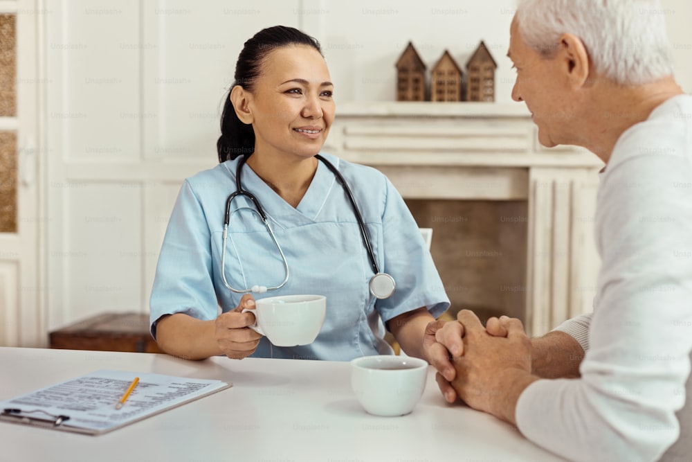 Pleasant drink. Cheerful delighted nice nurse holding a cup and having tea while talking to her elderly patient