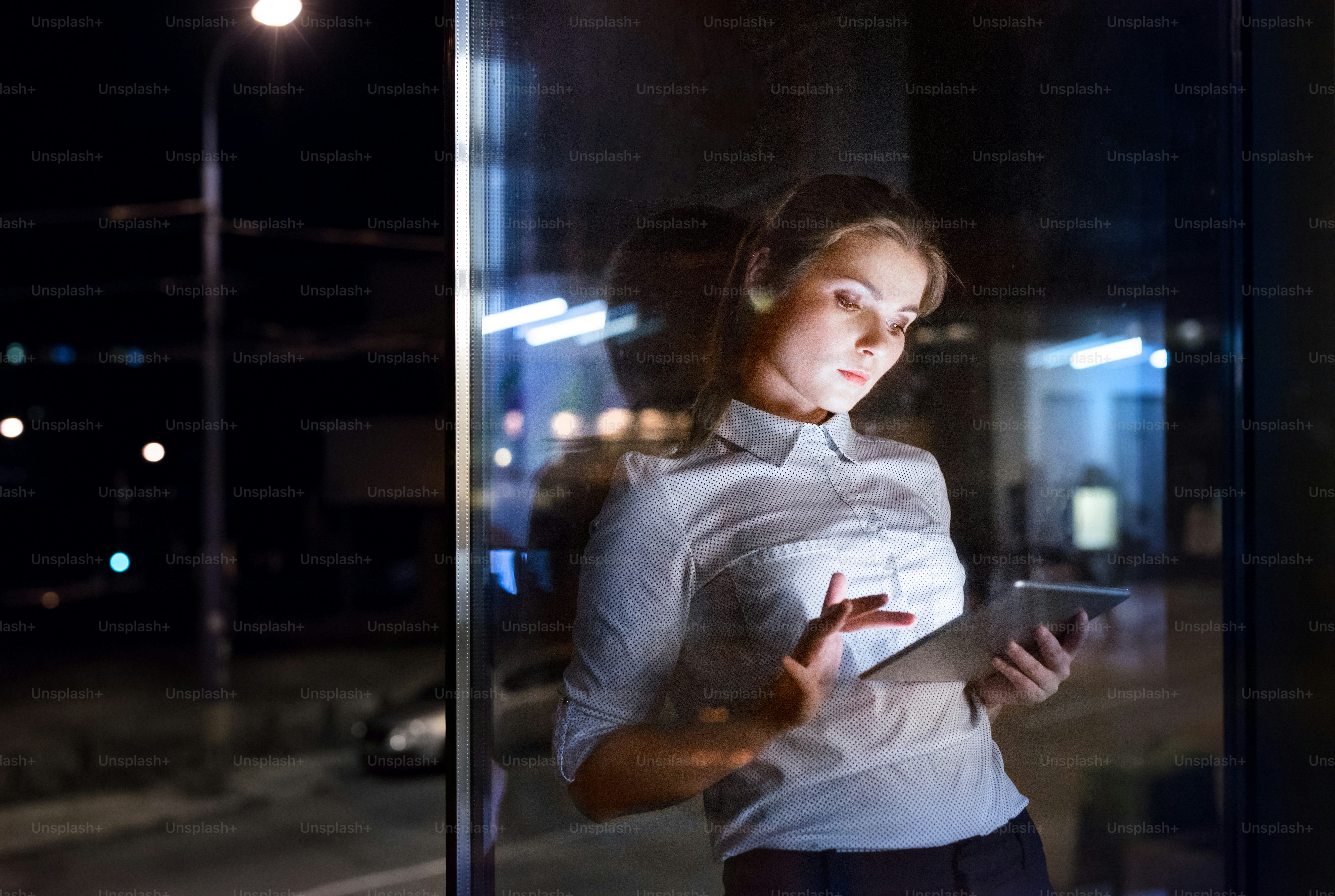 Beautiful young businesswoman with tablet in the office working late at night. Shot through glass.