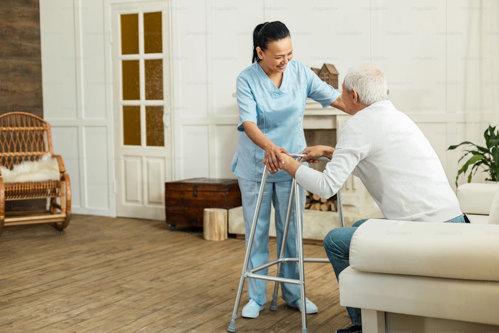 I will help you. Joyful delighted professional caregiver smiling to her patient and helping him to get up while doing her job