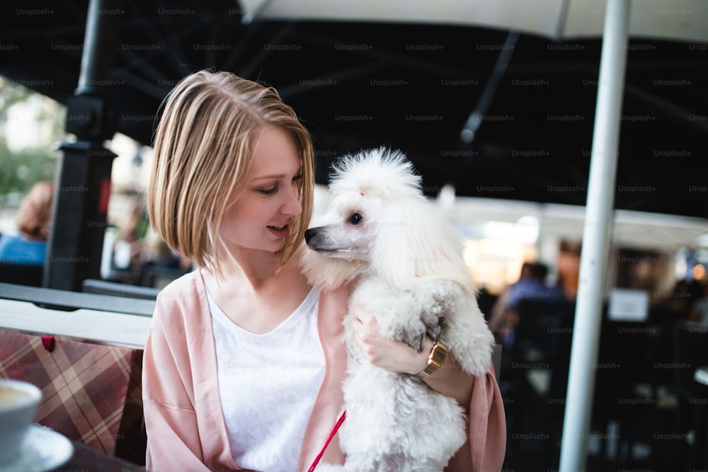Beautiful and fashionable young woman sitting in cafe bar with her gorgeous white dwarf poodle and looking at camera