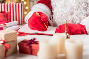 Close up of male hands in red gloves writing in notebook by pen. Santa Claus is sitting at desk near various presents