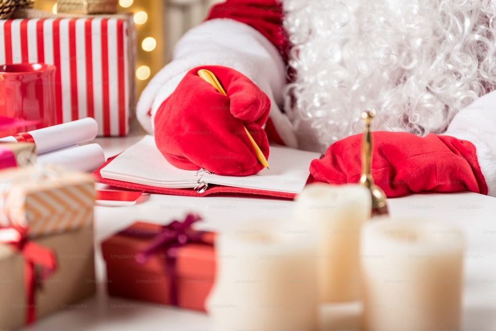 Primer plano de manos masculinas con guantes rojos escribiendo en cuaderno con bolígrafo. Papá Noel está sentado en el escritorio cerca de varios regalos