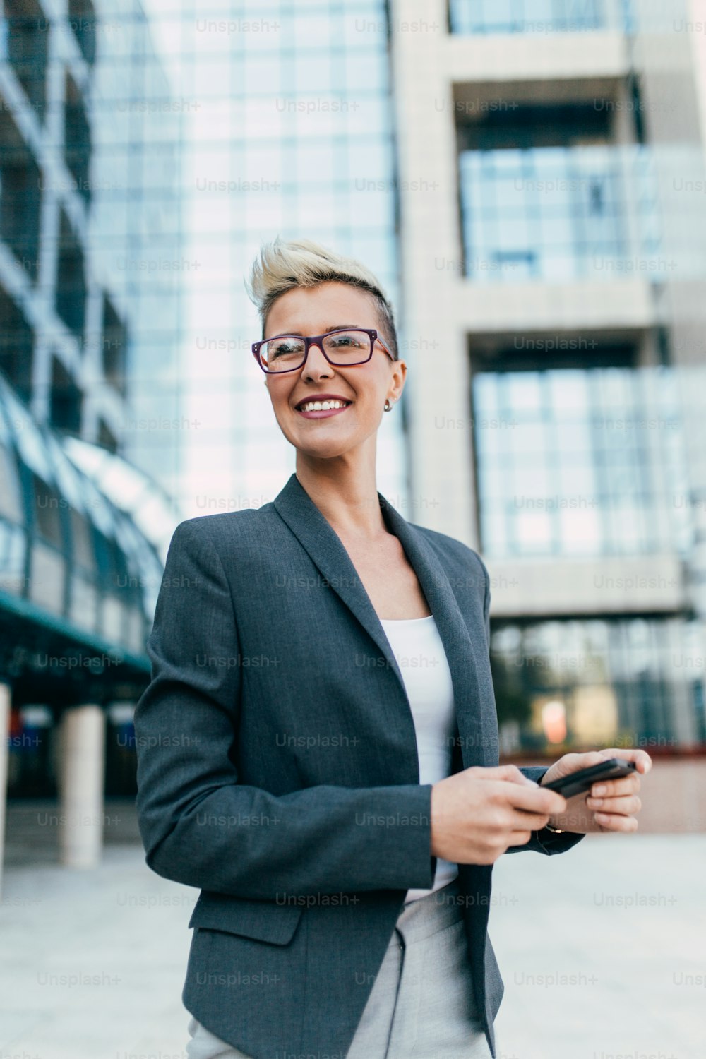 Jeune femme d’affaires heureuse debout devant un grand bâtiment moderne. Elle sourit et parle sur son téléphone portable.