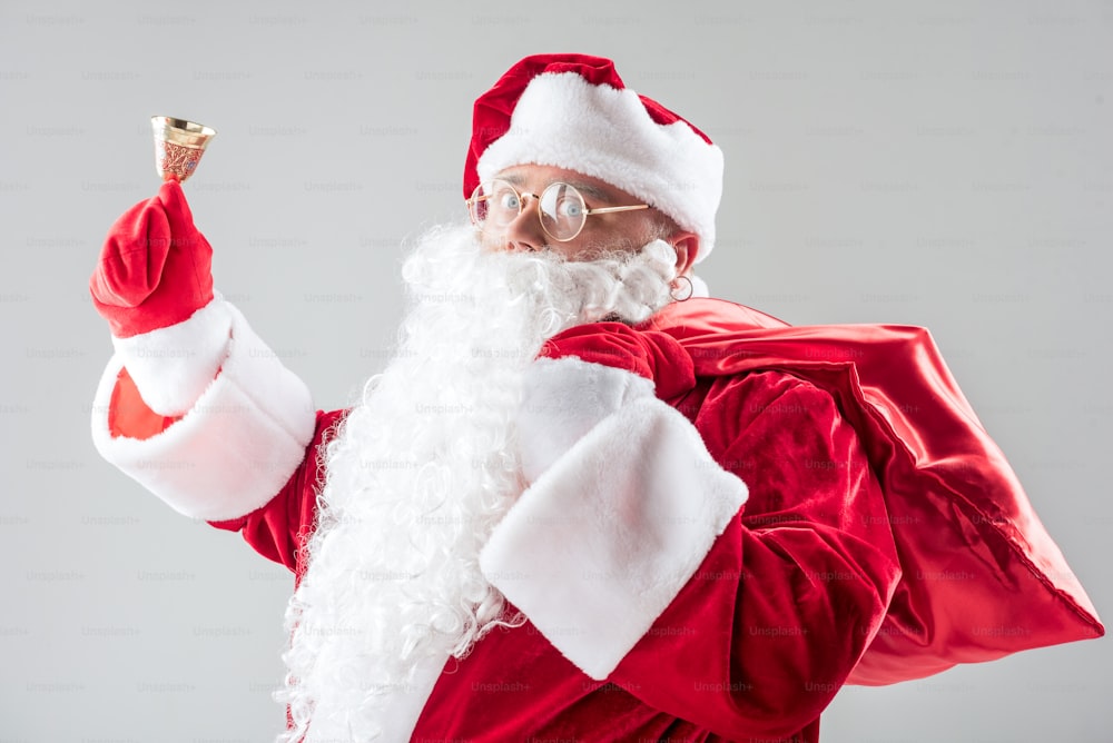 Schelle. Taillenporträt des aufgeregten Weihnachtsmanns, der die Glocke läutet, während er einen roten Sack hält. Isoliert auf grauem Hintergrund