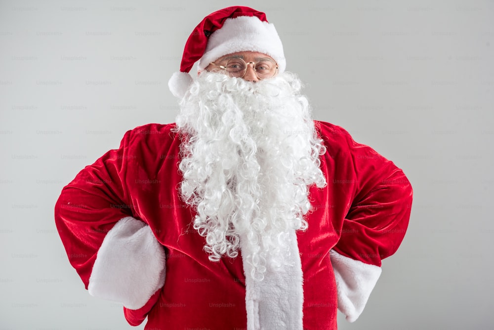 Waist up portrait of proud bearded Santa Claus standing with arms akimbo. He is looking at camera and smiling. Isolated on grey background