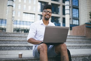 Young attractive Afro American businessman sitting on stairs, drinking coffee in front of huge modern building and doing some work on laptop.