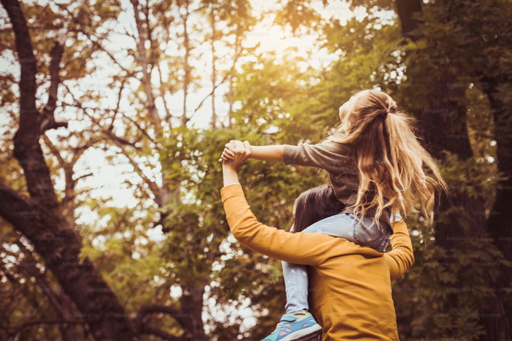 Mother and daughter outdoors in a meadow. Mother carrying her daughter on shoulder. From back.