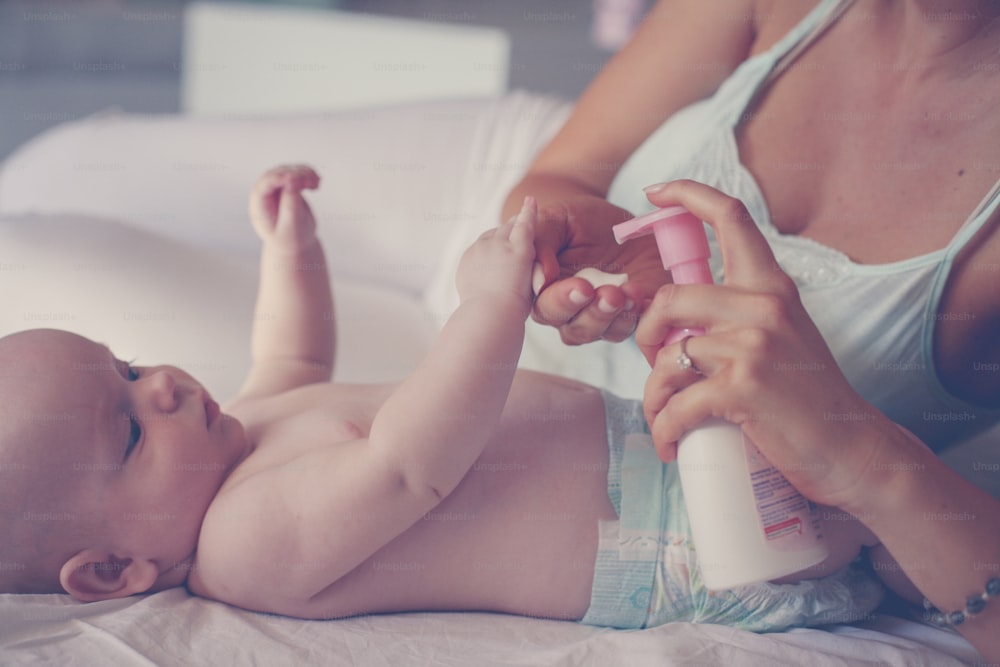 Mother with her baby by at home. Mother uses a body cream, and putting of a baby.