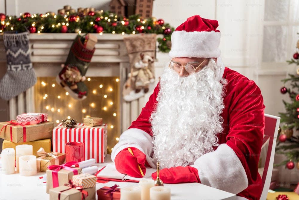 Ritratto di Babbo Natale serio sta prendendo appunti sul suo taccuino. È seduto alla scrivania vicino ai regali di Natale a casa