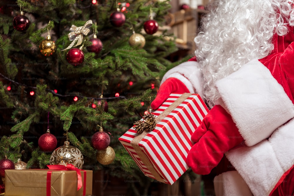 Close up das mãos do Papai Noel colocando presente de Natal sob o abeto. Ele está usando luvas vermelhas